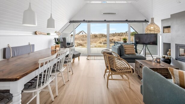 A modern interior with a wooden dining table, white chairs, a sofa, and a large window offering a view of the outdoors.
