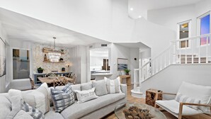 A spacious living room with a gray sofa, a glass coffee table, and a wooden side table. A staircase with white railings leads to an upper level. - Backyard Oasis w/ Fire Table + Walk to Salt Creek Beach | Dana Point Dreams (Dana Point)