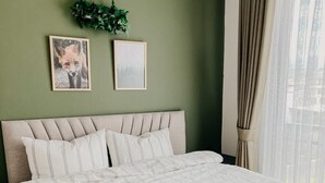 Cama bem arrumada com manta verde e roupa de cama listrada, cabeceira com ripas verticais, duas imagens emolduradas na parede e uma janela com cortinas leves.
