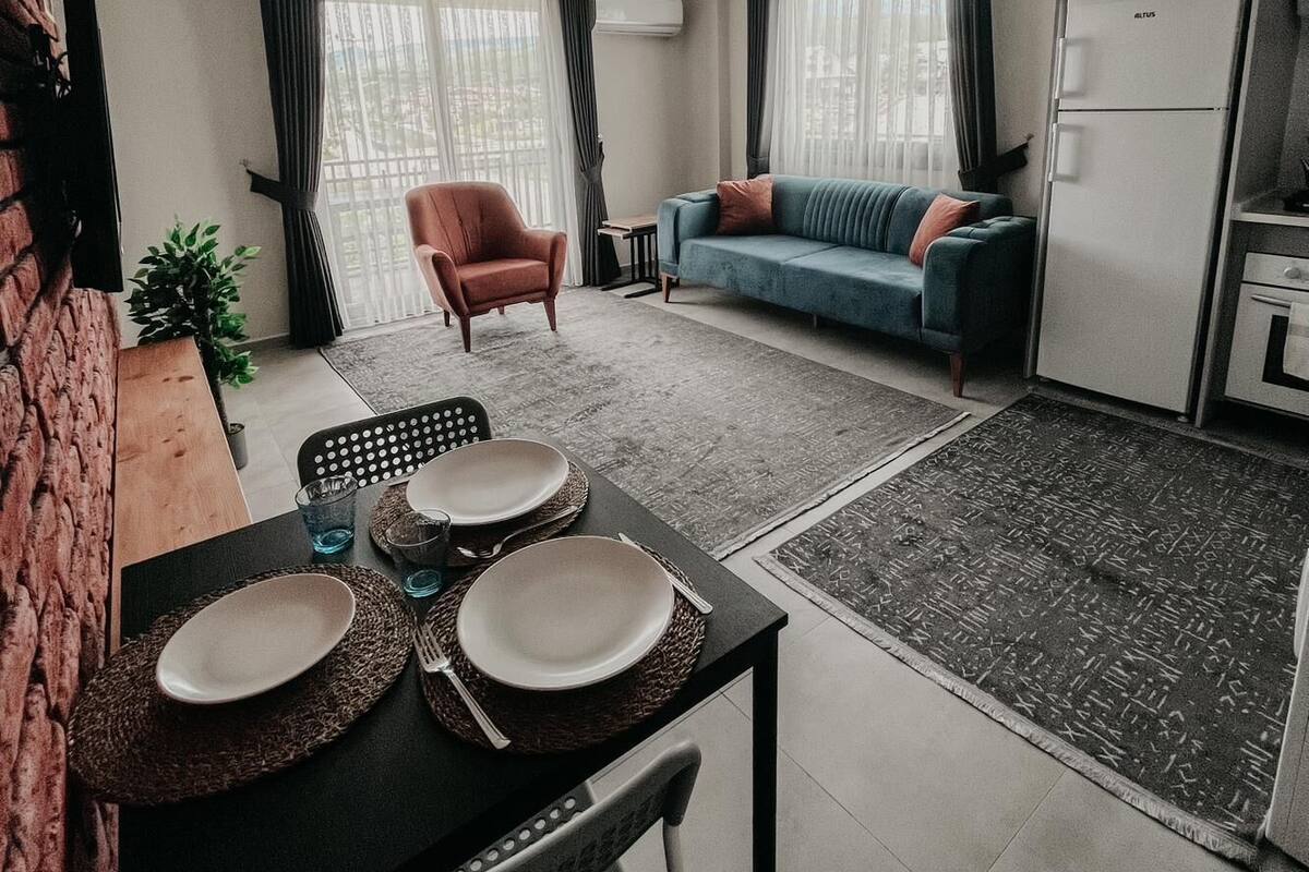 Una sala de estar moderna con comedor, un sofá, una silla y una zona de cocina con refrigerador.