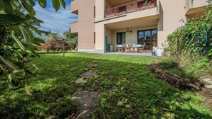 Un edificio residencial con balcón, un pequeño patio con mesa y sillas, y un jardín con césped y piedras que forman un camino.