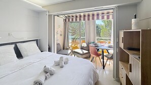 A hotel room with a large bed, a balcony with a table and chairs, and a wooden bookshelf. - Studio Coco`B, 4p, A/C, Wi-Fi, beach 500m, parking (Fréjus)