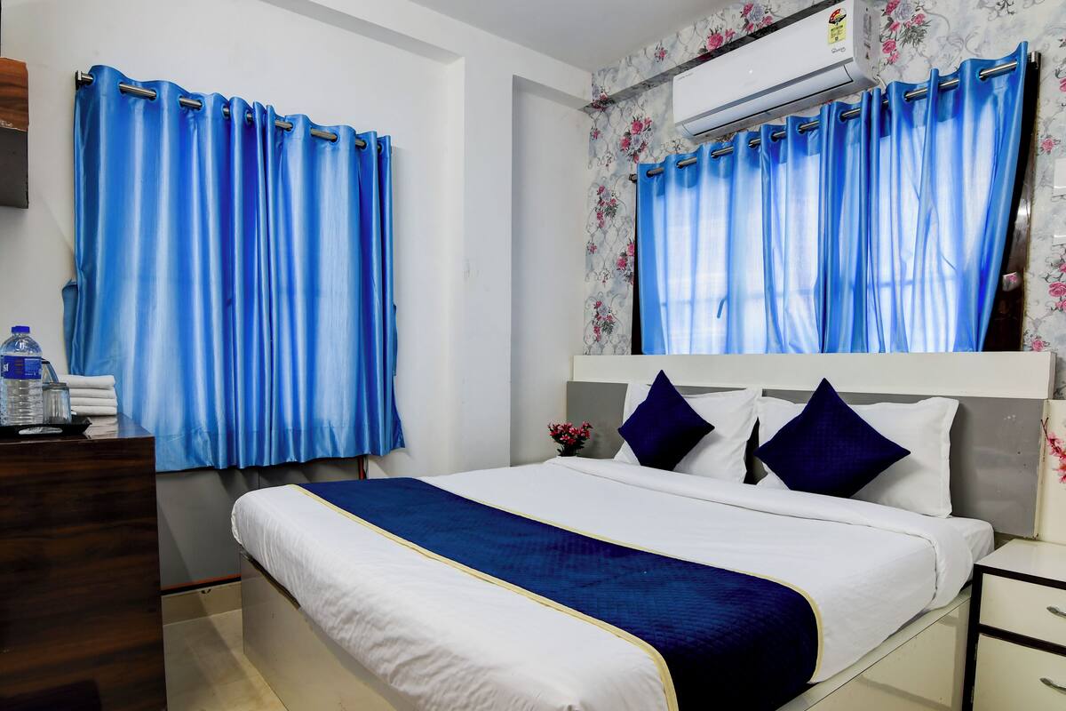 Une chambre d’hôtel avec un lit, des rideaux, un bureau, une bouteille d’eau et un vase à fleurs.