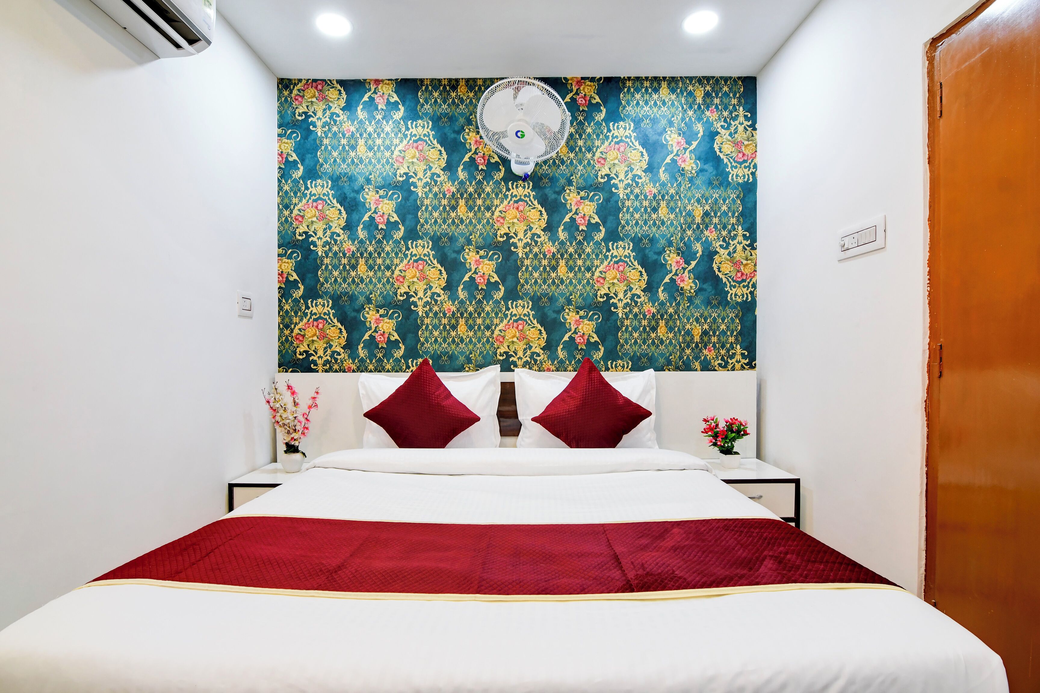 A hotel room with a bed, a wall-mounted air conditioner, a ceiling fan, and a wooden door.