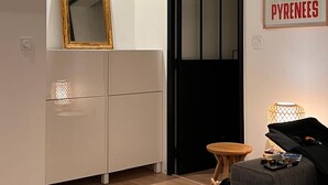 A modern living space with a wooden ceiling, a white cabinet, a mirror, a framed poster, a small table with a lamp, and a sofa with a board game. - Lovely, warm, fully renovated apartment in the center of the village (Ax-les-Thermes)