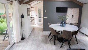Et moderne spiseområde med et rundt bord, seks stole og en vase med blomster. Der er en skydedør af glas, der fører til et udendørs område.