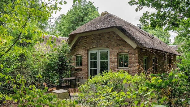 A rustic brick house with a thatched roof, surrounded by lush greenery and a small outdoor seating area. - Romantic Loft w/ Garden Terrace (Ermelo)