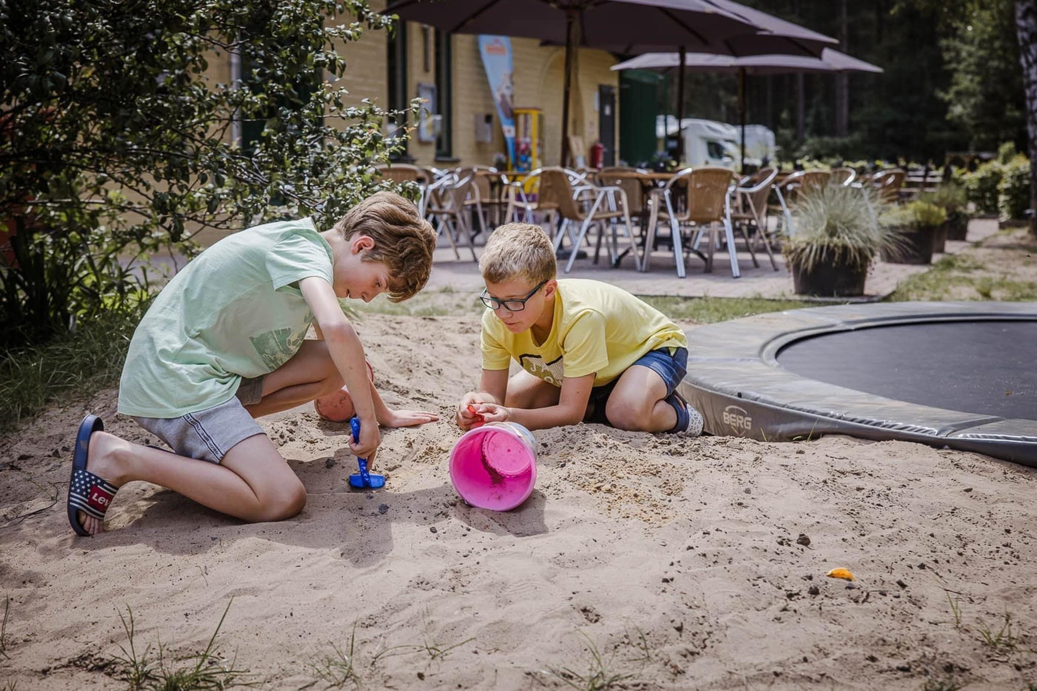 Two children playing with sand near a trampoline and outdoor seating area.