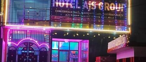 Un edificio con señalización iluminada para Hotel AJS Group, que indica que ofrece salón de conferencias, salón de bodas y habitaciones con aire acondicionado.