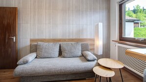 Ein modernes Zimmer mit grauem Sofa, zwei Holzstühlen, einer Holztür und einem Fenster mit Blick auf Grünflächen.