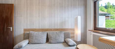 A modern room with a grey sofa, two wooden chairs, a wooden door, and a window with a view of greenery.