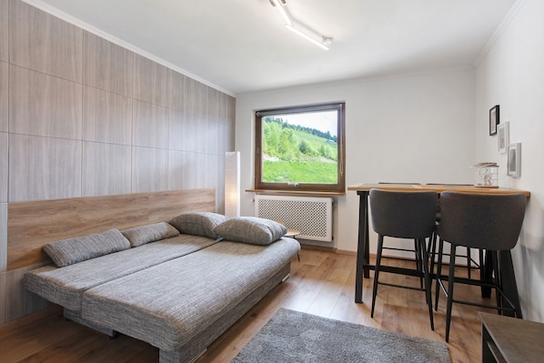 A bedroom with a bed, a desk, two chairs, a radiator, and a window with a view of greenery.