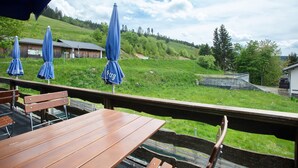 Eine Holzterrasse mit Bänken und blauen Sonnenschirmen, mit Blick auf einen grasbewachsenen Hügel und ein Gebäude.