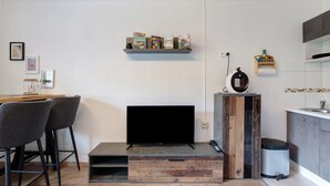 Ein modernes Wohnzimmer mit Flachbildfernseher, einem Holztisch, zwei Sesseln, einer kleinen Bücherregal und einer Küchenzeile mit Spüle und Schränken.