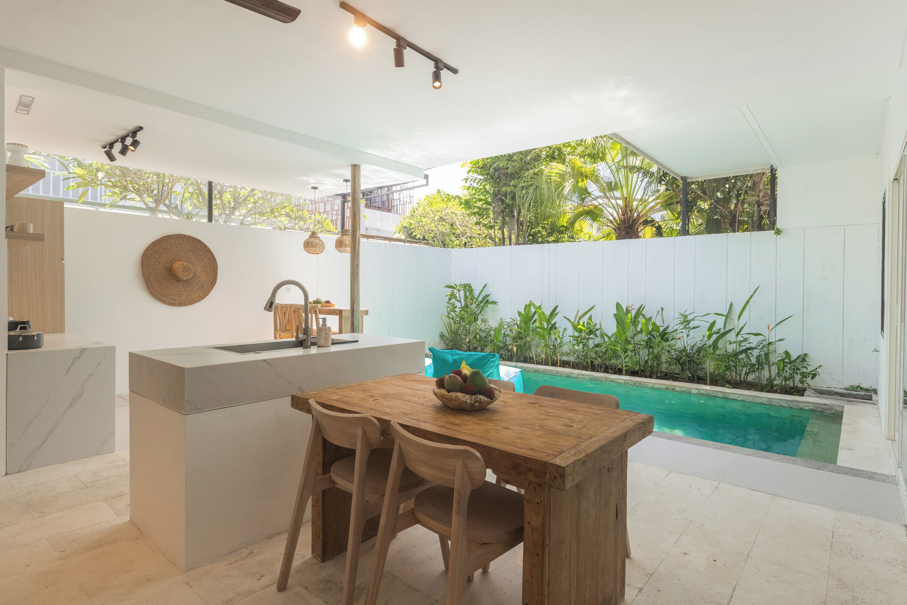 Una cocina moderna con una mesa y sillas de madera, una isla empotrada con fregadero y vistas a una piscina rodeada de vegetación.