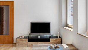 A modern living room with a flat-screen TV on a glass-top coffee table, wooden cabinets, and a window with a view.