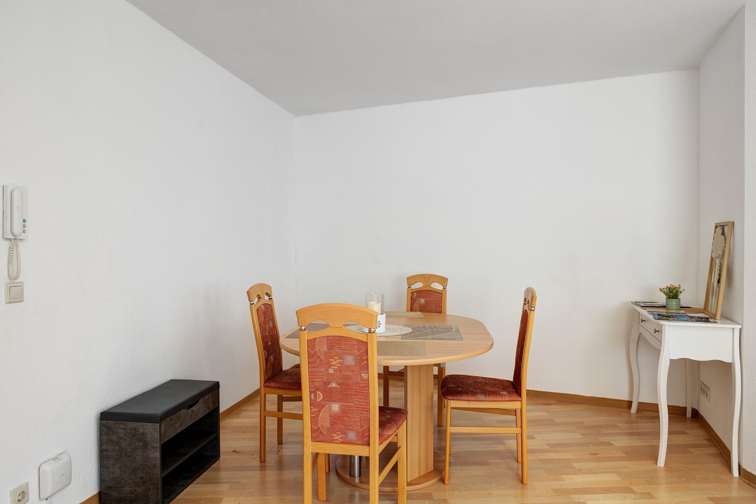 Een eetgedeelte met een ronde tafel, vier houten stoelen, een klein wit bureau met spiegel en een bank.