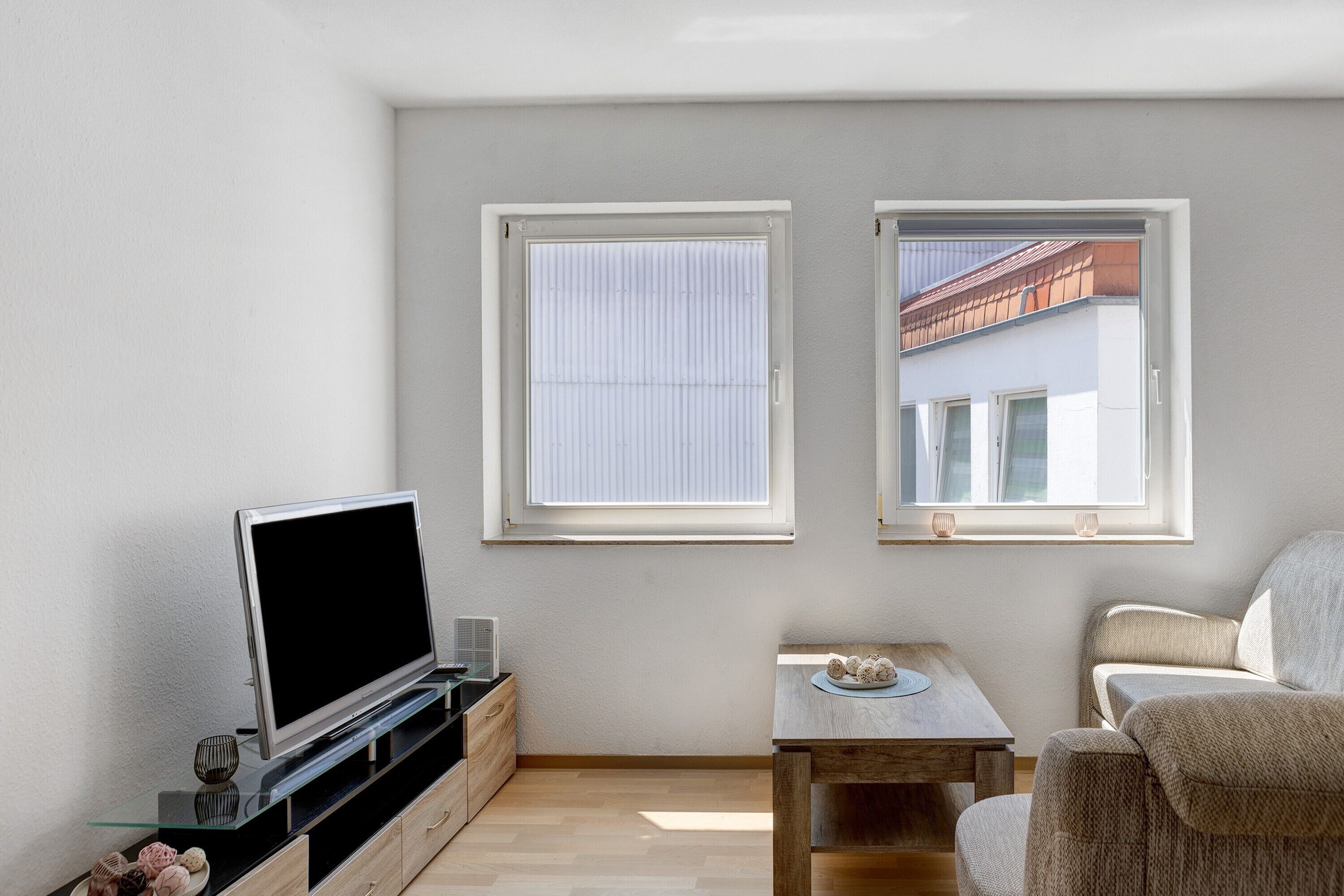 A modern living room with a flat-screen TV, a small table, and a sofa. There are two windows providing natural light.