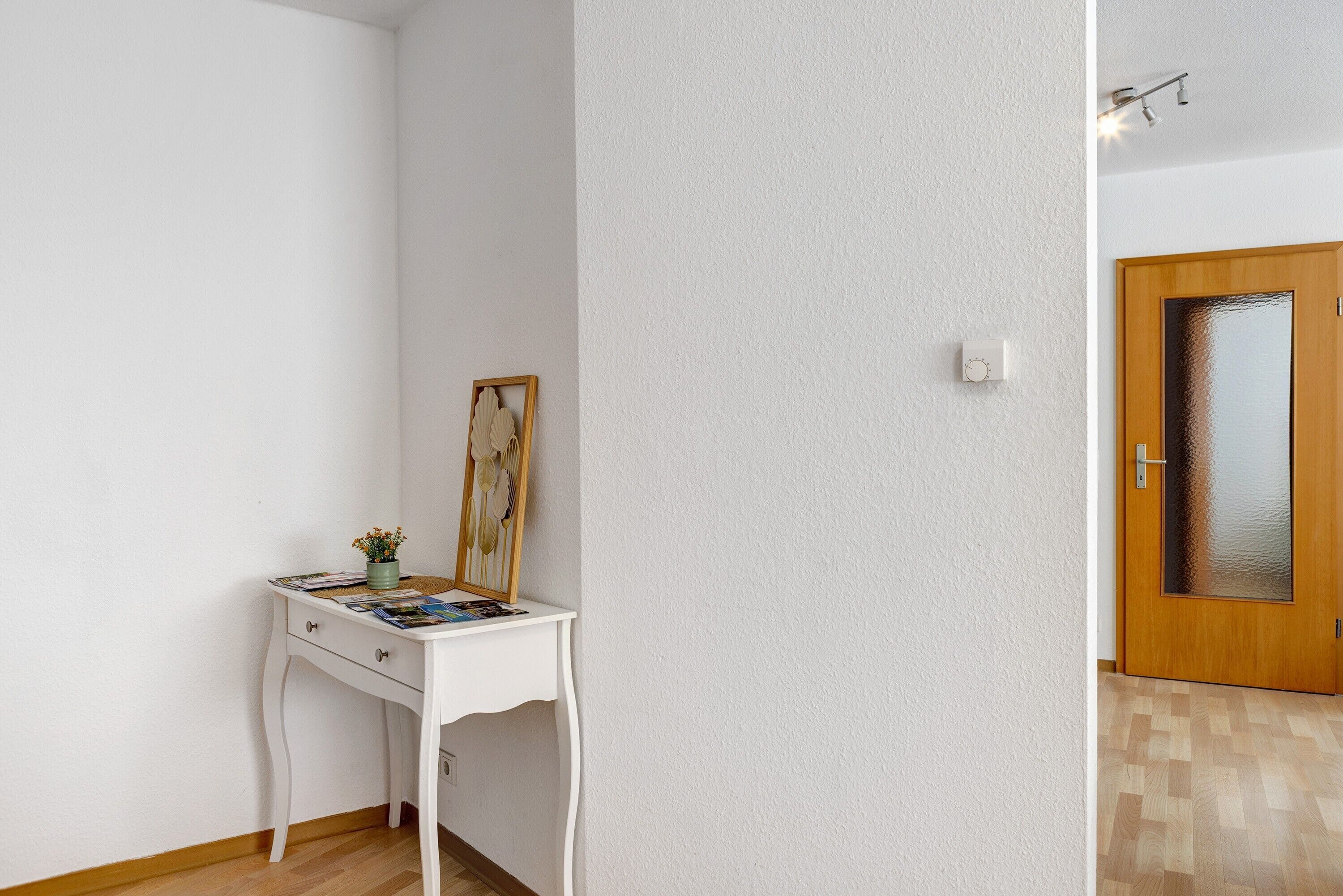 En vit vägg med en vit dörr, ett träkarm, och ett litet vitt bord med en växt och en inramad bild.