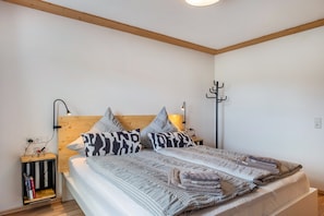 Un dormitorio con cabecera de madera, una mesita de noche con lámpara, una estantería y una cama con ropa de cama estampada.
