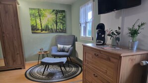 A room with a wooden cabinet, a grey armchair, a small round rug, a television on the wall, and a window with curtains.
