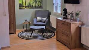 A room with a wooden cabinet, a grey armchair with a forest-themed pillow, a small round table with a decorative item, a radiator, and a television mounted on the wall.