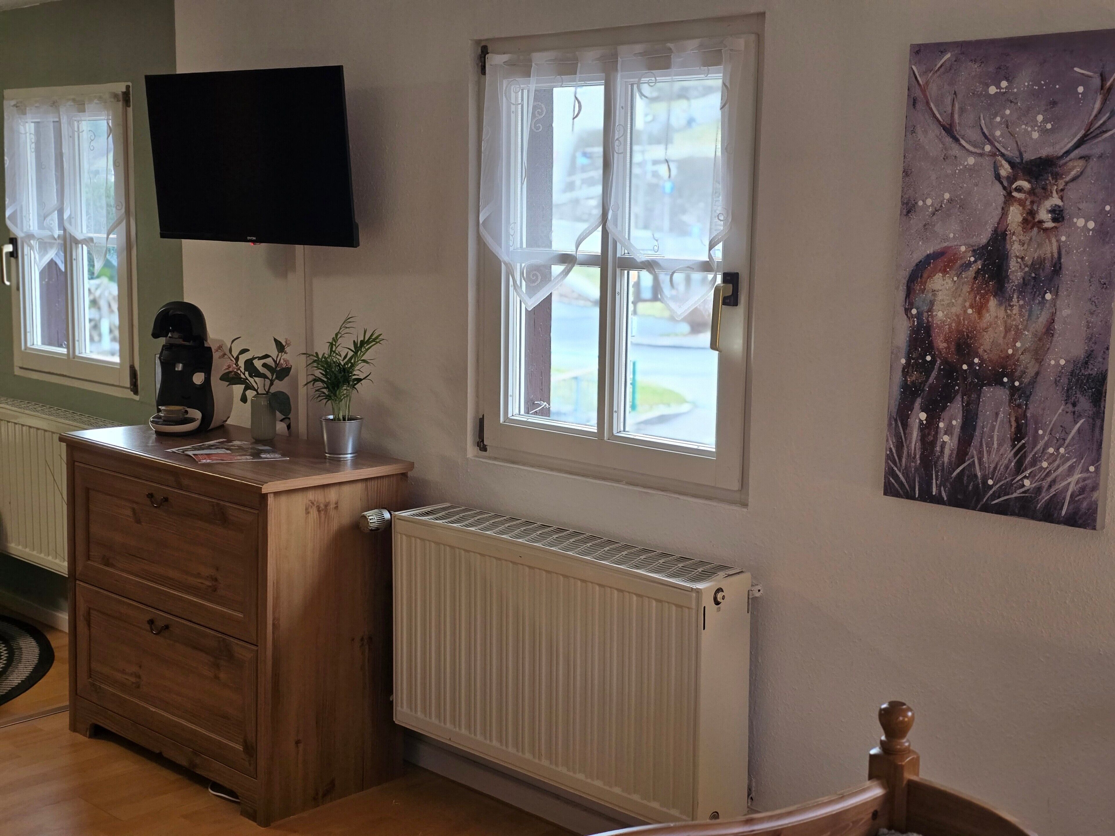 Una habitación con un aparador de madera, un radiador, un televisor montado en la pared y una ventana con cortinas.