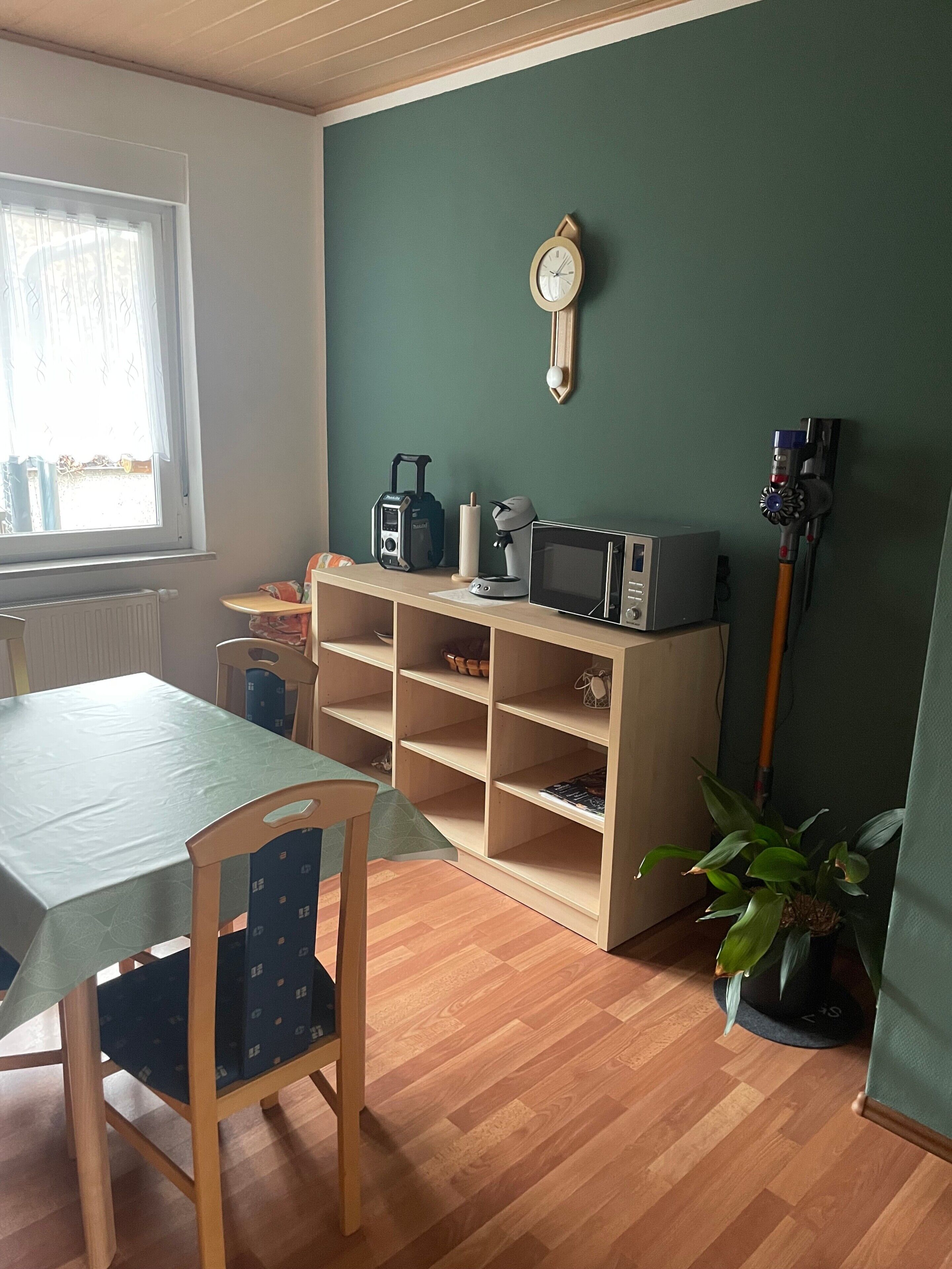 A room with a dining table, chairs, a bookshelf, a clock, a coffee maker, a microwave, and a vacuum cleaner.