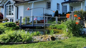 Une grande maison blanche avec de nombreuses fenêtres, un jardin où poussent différentes plantes et fleurs, et un petit pont.