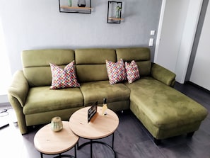 Un canapé d’angle vert agrémenté de coussins à motifs, deux petites tables rondes et une petite étagère ornée d’objets décoratifs, le tout fixé au mur.