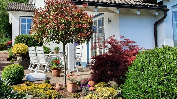 Une maison blanche avec un toit en tuiles, une terrasse avec des meubles d’extérieur et diverses plantes en pot.