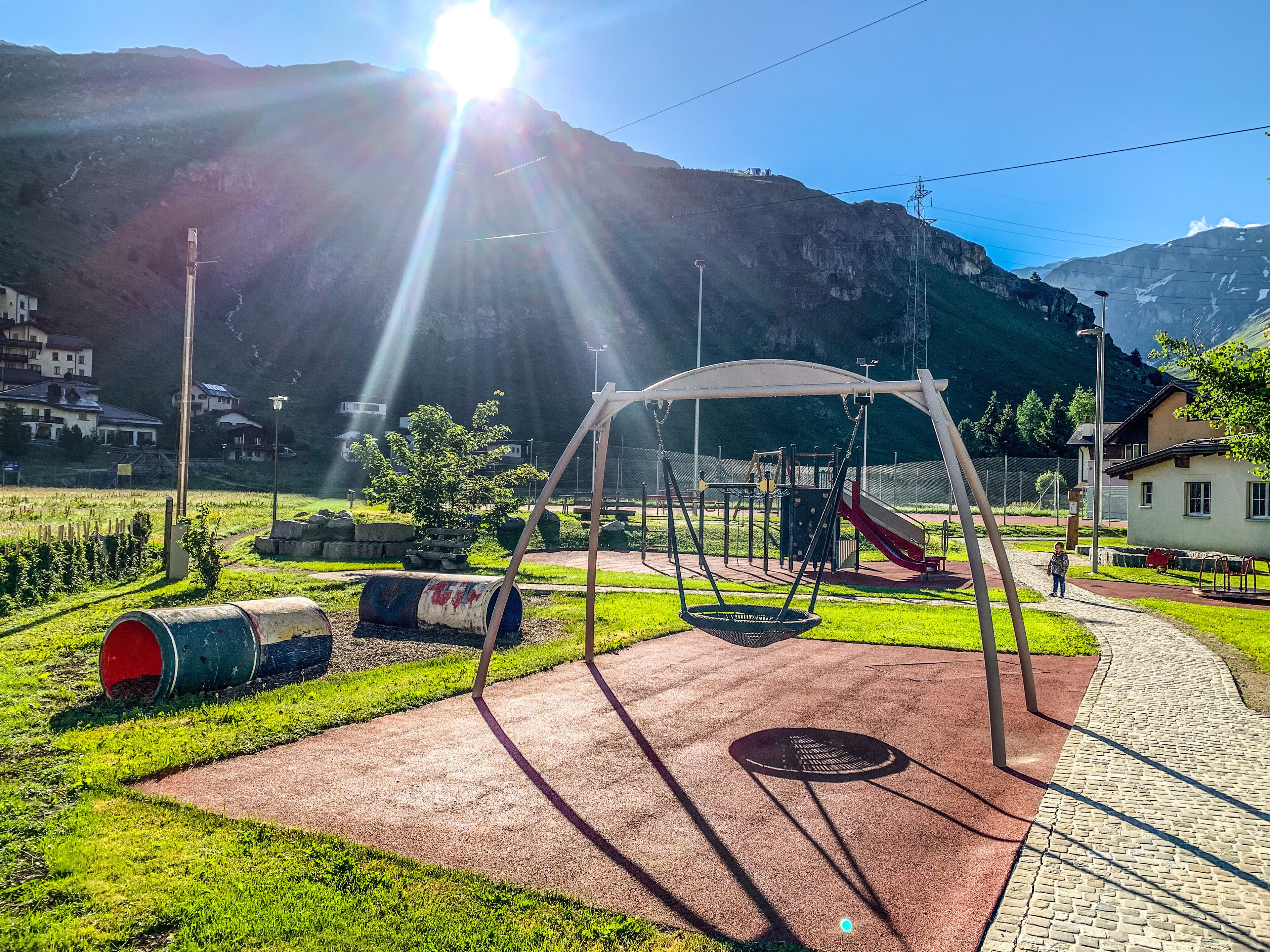 Un terrain de jeux avec des balançoires et un toboggan, entouré d’une zone pavée et de verdure, avec des maisons et des montagnes en arrière-plan.