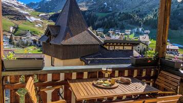 Une terrasse en bois avec une table dressée pour le repas, offrant une vue sur un village aux bâtiments traditionnels et sur un paysage montagneux en arrière-plan.