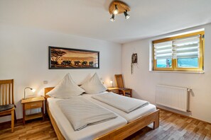 Ein Schlafzimmer mit Bett, Nachttischen, einem Sessel, einem Schreibtisch, einem Fenster mit Jalousien, einer Heizung und einem Wandbild.