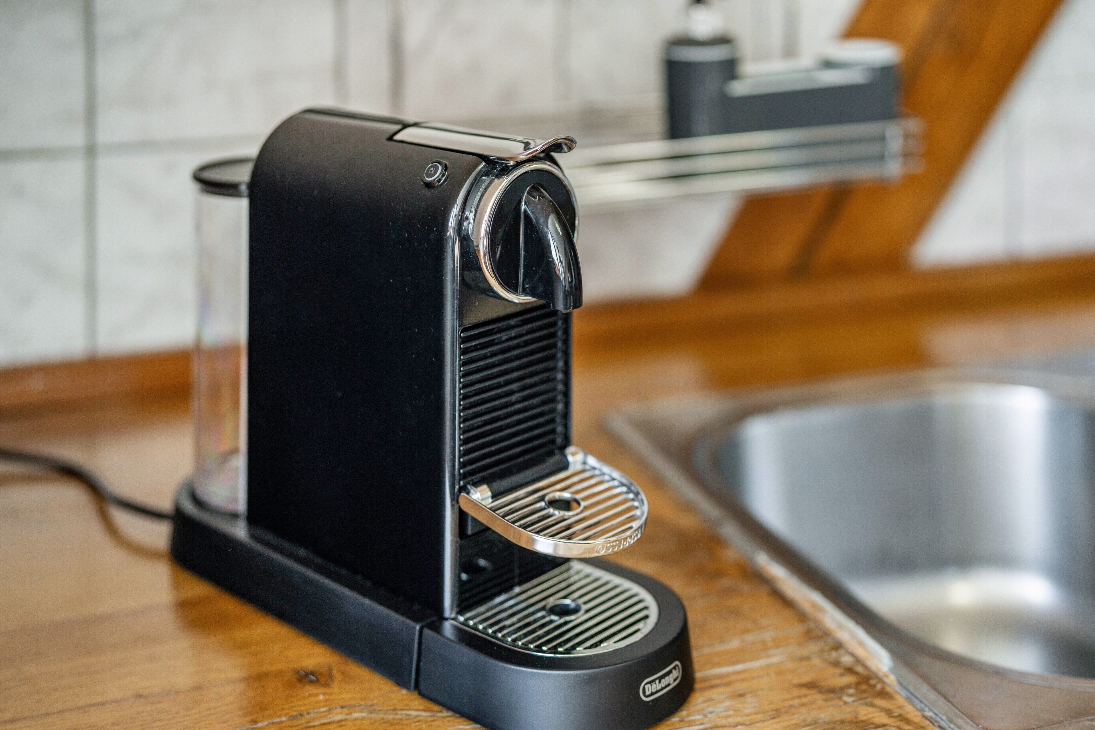 A black coffee machine on a wooden countertop with a stainless steel sink in the background.