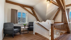 Un dormitorio tipo loft con techo inclinado, una cama con cabecera de madera, un sillón gris, una mesita redonda con una planta y una ventana con cortinas.