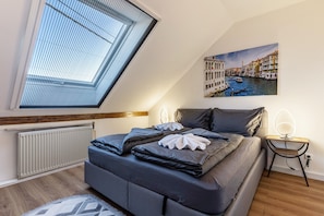 A bedroom with a skylight, a bed with dark bedding, a painting of a canal, and a bedside table with a lamp.