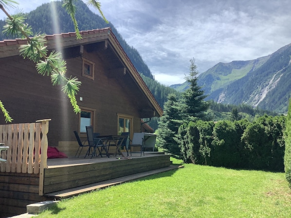 A wooden cabin with a deck, surrounded by lush greenery and mountains.