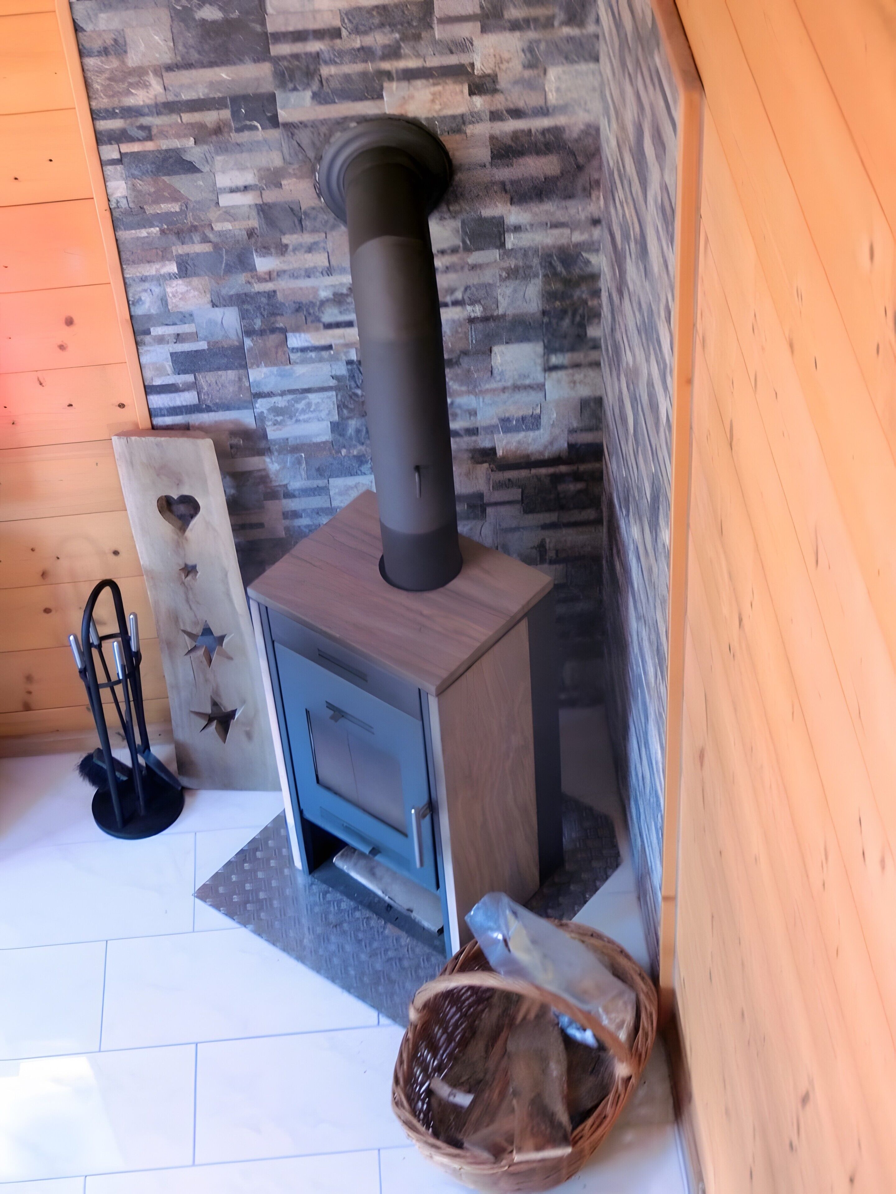 A wood-burning stove, a metal rack, and a woven basket in a room with a stone wall and wooden flooring.