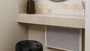 Une salle de bain avec un miroir rond, un vase contenant de l’herbe des pampas, un tabouret noir à motifs et un mur recouvert de carreaux.
