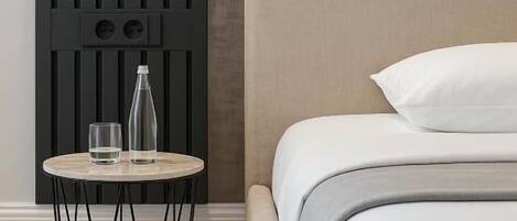 A neatly made bed with a white pillow and a beige headboard, a round side table with a clear glass and a bottle, and a black wall with vertical slats and a light fixture.