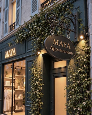Un immeuble avec une enseigne indiquant « Appartements MAYA » et une vitrine où sont exposés des objets soigneusement disposés.