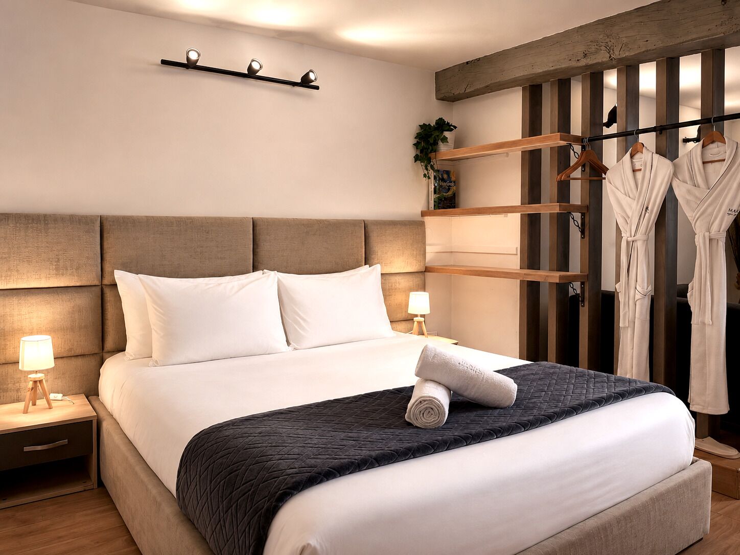 Ein ordentlich bezogenes Bett mit weißer Bettwäsche, einer dunkel gesteppten Decke und einem aufgerollten Handtuch. Zwei Nachttischlampen, ein Holznachttisch und eine wandmontierte Leuchte sind ebenfalls vorhanden.