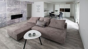 Un salon moderne avec un canapé d’angle, une petite table ronde agrémentée de deux vases décoratifs, une cuisine en arrière-plan et un mur orné d’un motif.