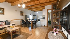 Een gezellige woonkamer met een bank, eettafel en stoelen, houten balken in het plafond, een keuken op de achtergrond en een boekenkast met decoratieve objecten.