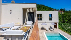 A modern outdoor pool area with lounge chairs and umbrellas, a wooden deck, and a view of a hillside.