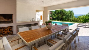 Un espace repas extérieur moderne, avec une table en bois et des chaises blanches, un barbecue intégré et une vue sur une piscine et la verdure.