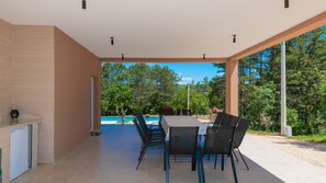 Um pátio externo com mesa e cadeiras, piscina e vista para árvores e céu.