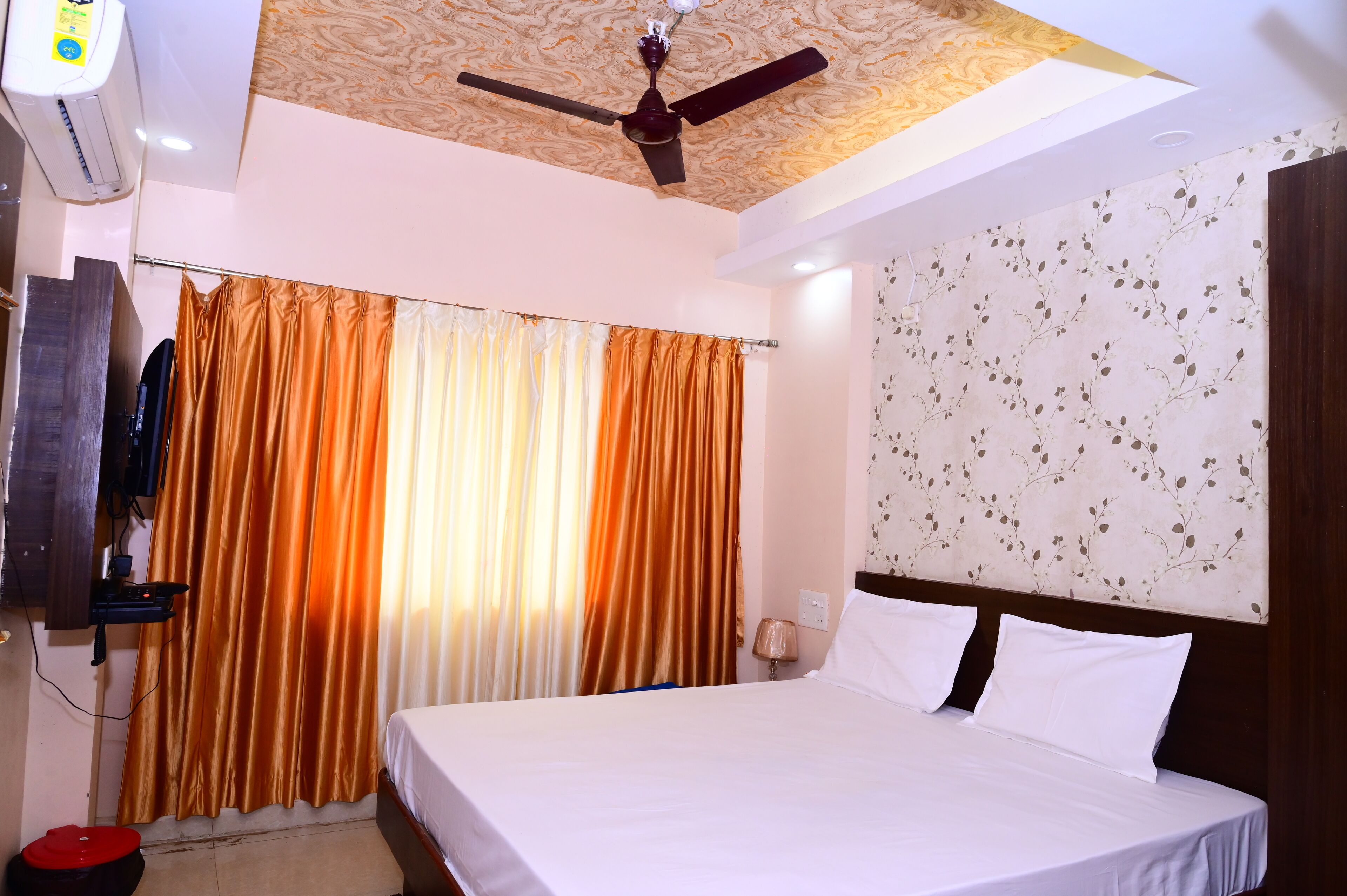 Habitación de hotel con cama, cortinas, ventilador de techo, aire acondicionado montado en la pared y cabecera estampada con motivos florales.