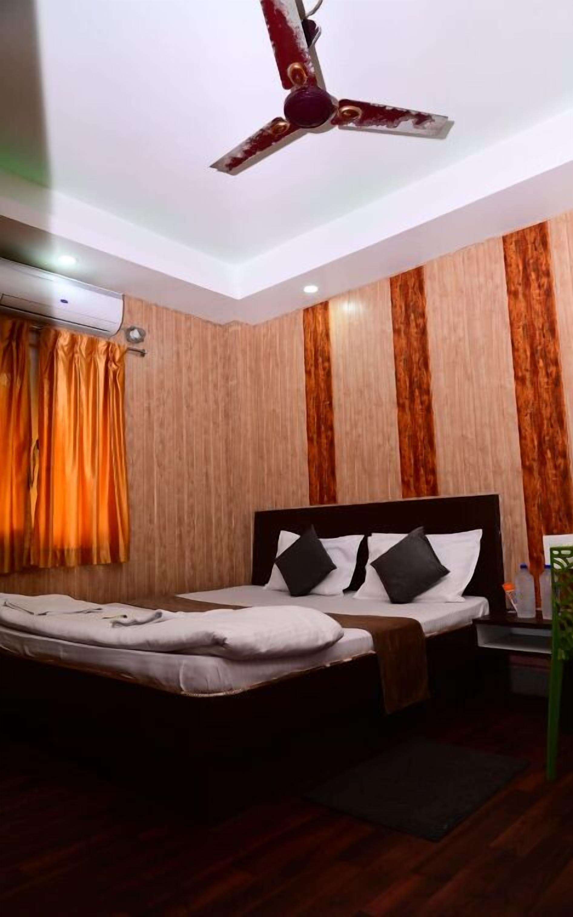 A hotel room with a bed, a ceiling fan, an air conditioning unit, wooden paneling, and a window with curtains.